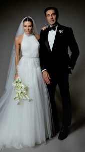 Esther Marques com seu vestido de noiva e um buque de flores, ao lado de seu marido, que usa um terno preto com camisa branca e gravata borboleta preta