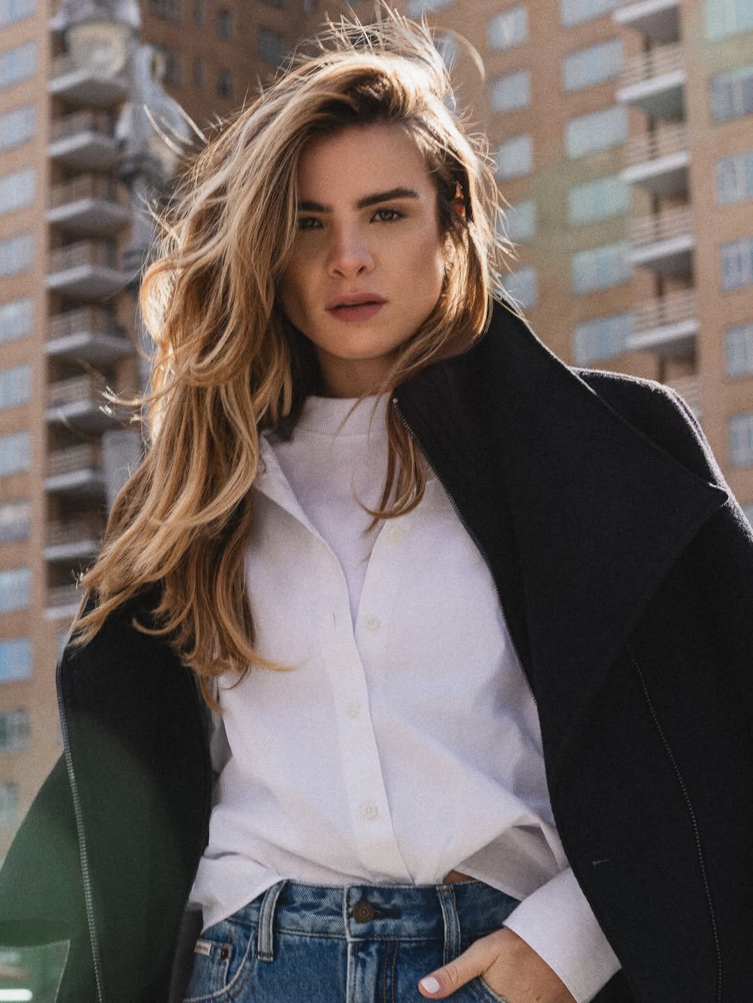 Cantora Giulia Be, branca de cabelos loiros e longos, posa para foto em cenário urbano usando uma cmaisa social branca, calças jeans e um casaco preto