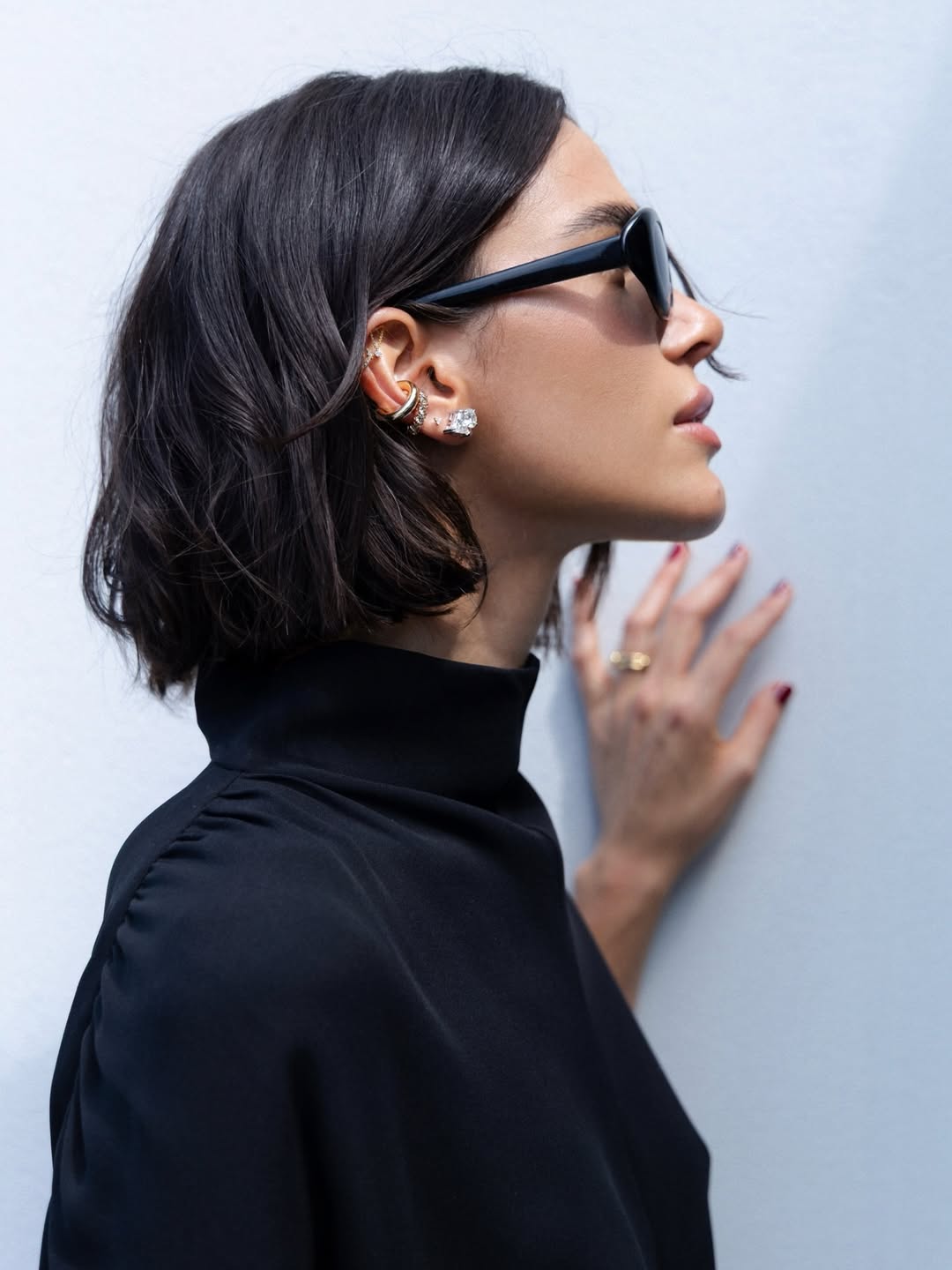 Bruna Marquezine, mulher, 30 anos, posa para foto com cabelos curtos e escuros, ólculo de sol, brincos brilhantes e camisa preta de gola rolê