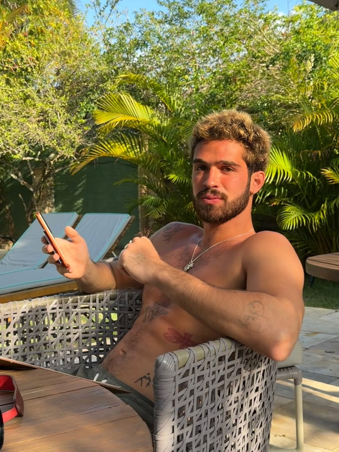 João Guilherme aparece sentado ao ar livre, sem camisa, usando o celular, em um ambiente com plantas e cadeiras de descanso ao fundo, sob luz natural