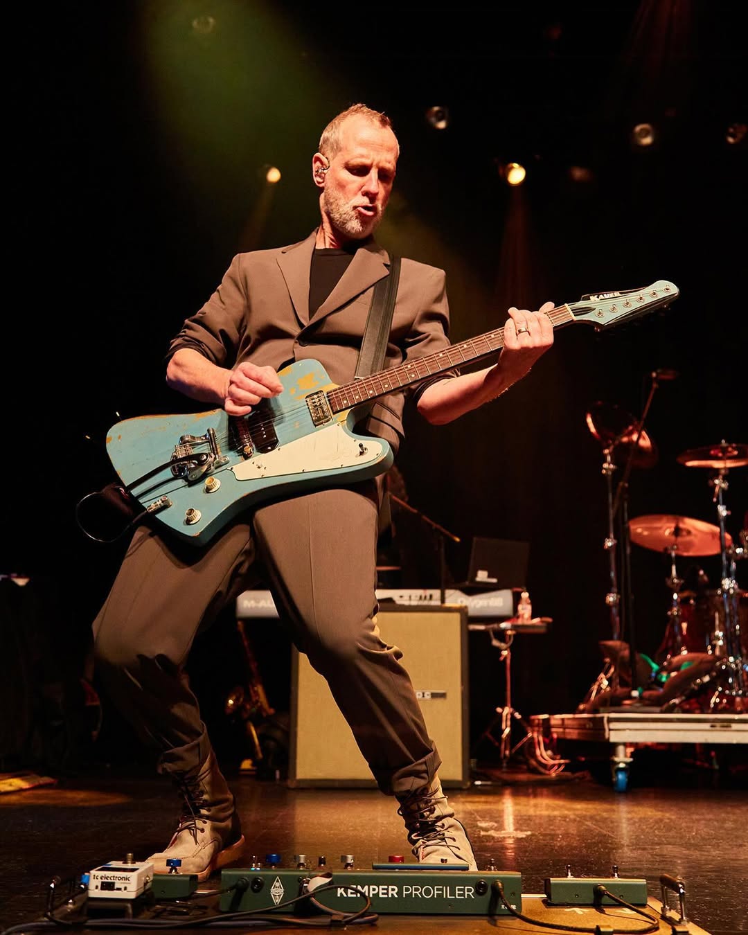 Uma fotografia de plano médio de Tom Dumont, guitarrista do No Doubt, tocando uma guitarra elétrica azul-petróleo desgastada no palco. Ele está em pé, com expressão focada, usando um terno cinza e botas. O palco está escuro com iluminação cênica. No chão, em frente a ele, há um pedalboard com equipamentos como um afinador branco e um processador Kemper verde. Ao fundo, desfocados, veem-se uma bateria e outros equipamentos de palco.