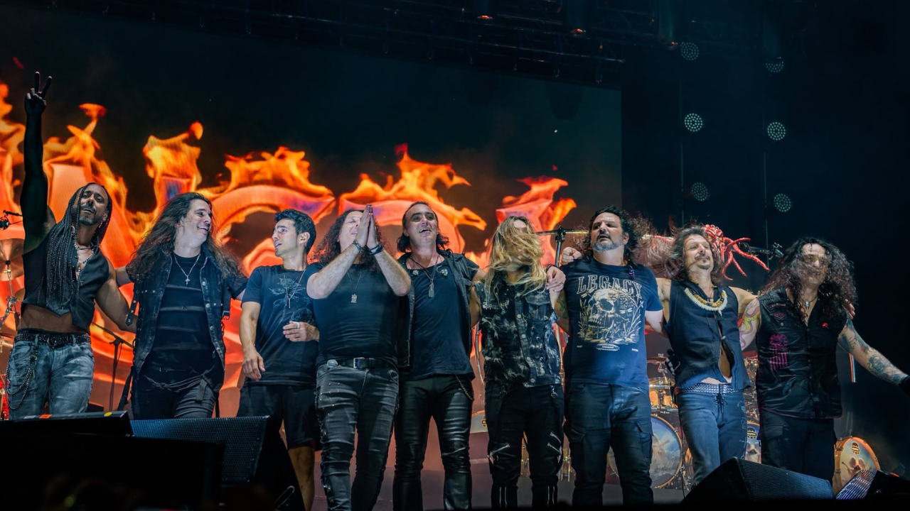 Fotografia de ângulo amplo e plano inteiro do palco principal de um festival de música. Nove músicos da banda Angra estão alinhados, abraçando-se com os braços entrelaçados ou em volta dos ombros, sorrindo e olhando para o público. O músico na extremidade esquerda tem cabelo longo encaracolado, barba, e ergue a mão direita com dois dedos (indicador e médio) estendidos. Os músicos vestem trajes clássicos de rock e metal, predominantemente pretos, incluindo calças de couro, jeans, camisetas de manga curta e coletes. Um músico no centro usa uma camiseta com uma estampa gráfica detalhada. Atrás deles, uma enorme tela de LED domina o fundo, exibindo gráficos vibrantes de fogo em tons de laranja quente e vermelho, possivelmente um logotipo estilizado da banda. Trusses de iluminação e spotlights estão visíveis acima da tela. Parte de uma bateria é visível atrás dos músicos à direita. A iluminação de palco é dramática e quente.
