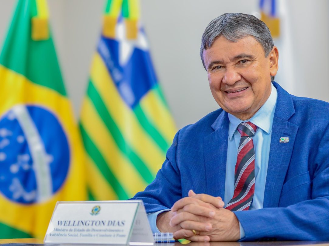 Wellington Dias aparece sentado à mesa, sorrindo e com as mãos apoiadas à frente do corpo. Ao fundo, as bandeiras do Brasil e do Piauí aparecem desfocadas