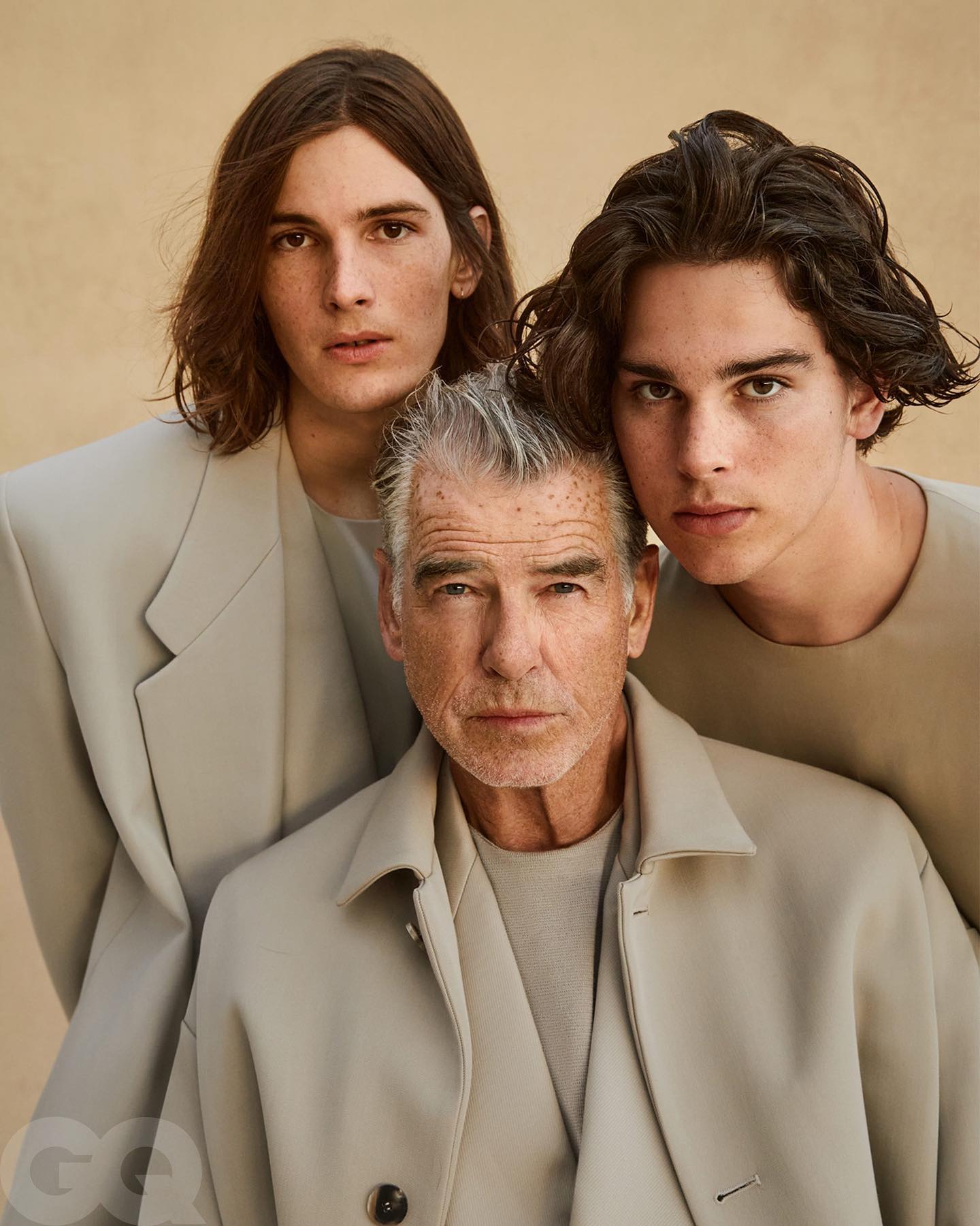 Pierce Brosnan aparece ao centro, usando roupas claras, ao lado dos filhos Dylan e Paris Brosnan, que estão atrás dele em um retrato posado. Os dois filhos do ator têm cabelos castanhos e vestem tons neutros.