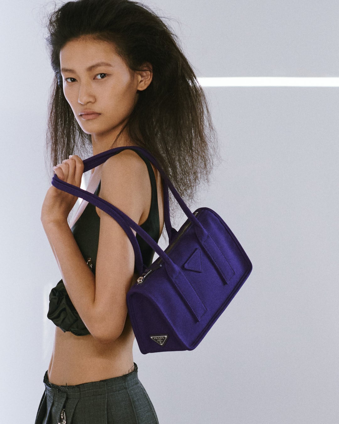 Fotografia de moda com uma modelo asiática de perfil, a segurar ao ombro uma mala retangular roxa de formato estruturado da Prada. A modelo exibe um cabelo escuro volumoso com textura frisada, veste um top preto curto e calças cinzentas, sob um fundo branco minimalista com uma linha luminosa horizontal.