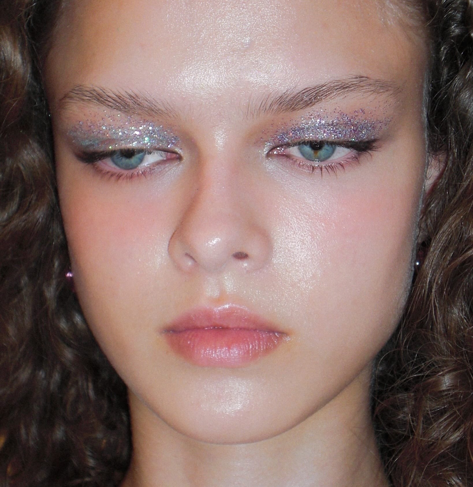 Close do rosto de uma modelo com maquiagem iluminada e olhos cobertos por glitter prateado, criando um efeito de sereia.