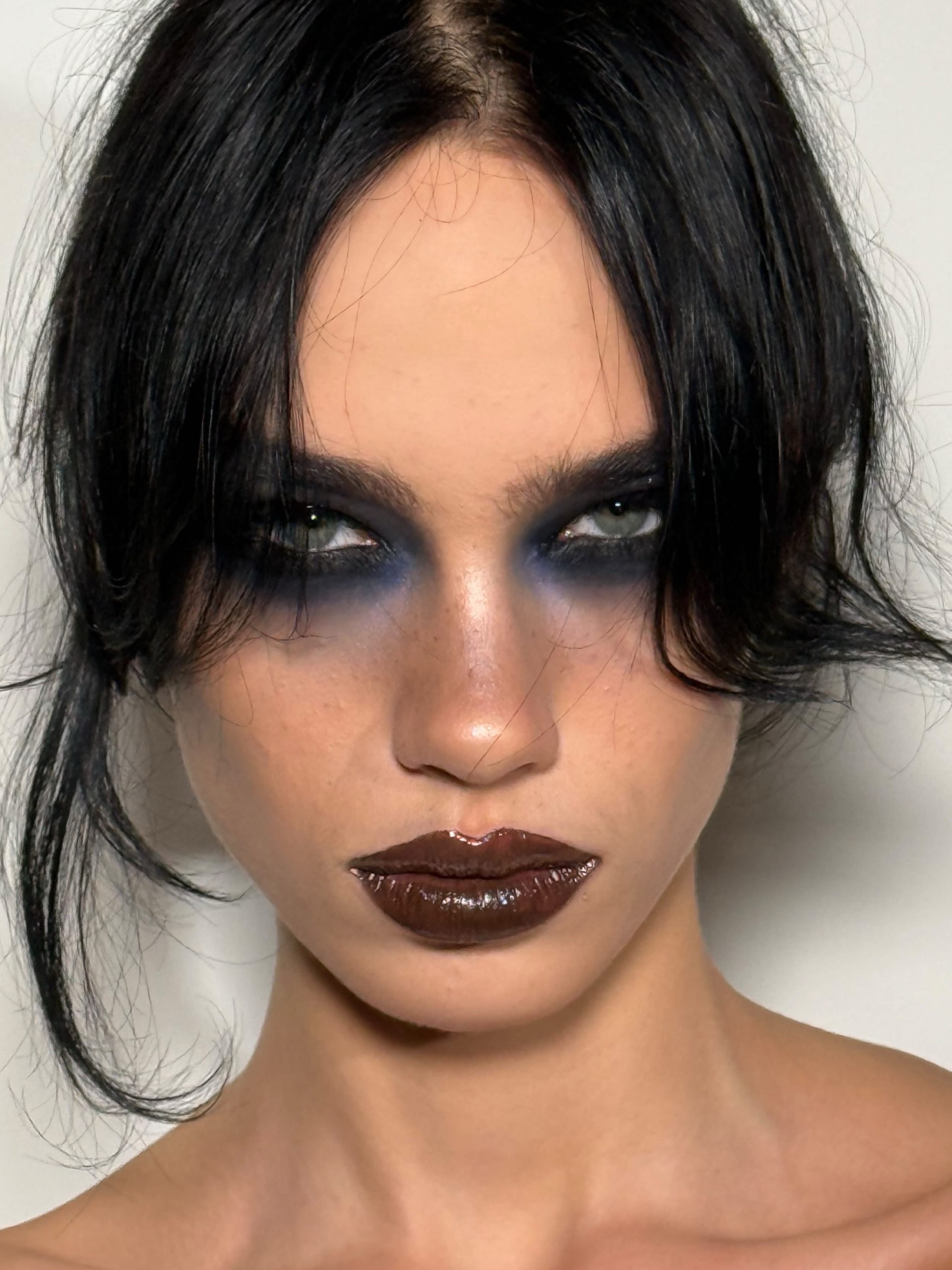 Close de uma modelo com maquiagem marcante, olhos esfumados em preto e azul e lábios em tom marrom com acabamento glossy.