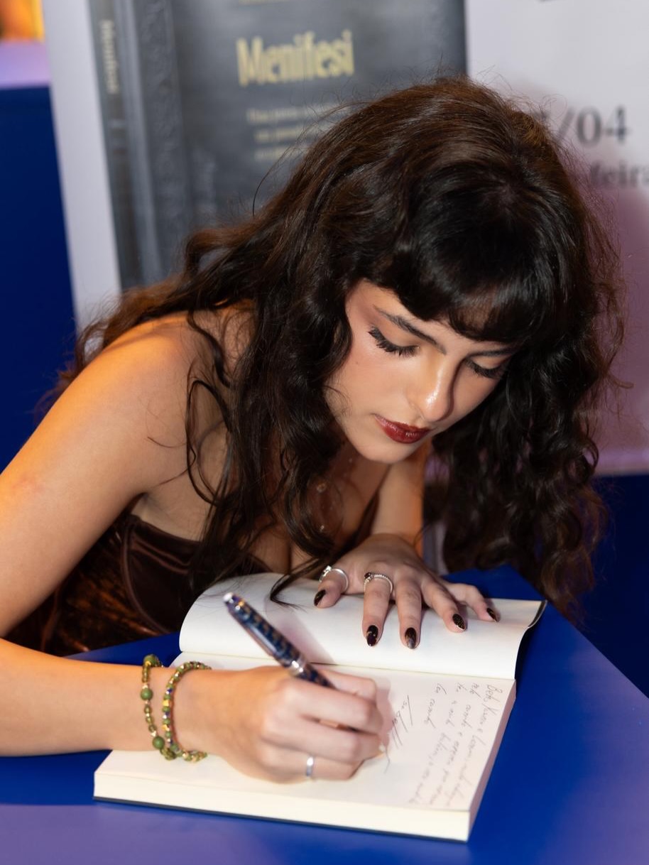 Isabella Arantes, branca, cabelos escuros, blusa vinho e calça jeans na sessão de autógrafos de seu livro