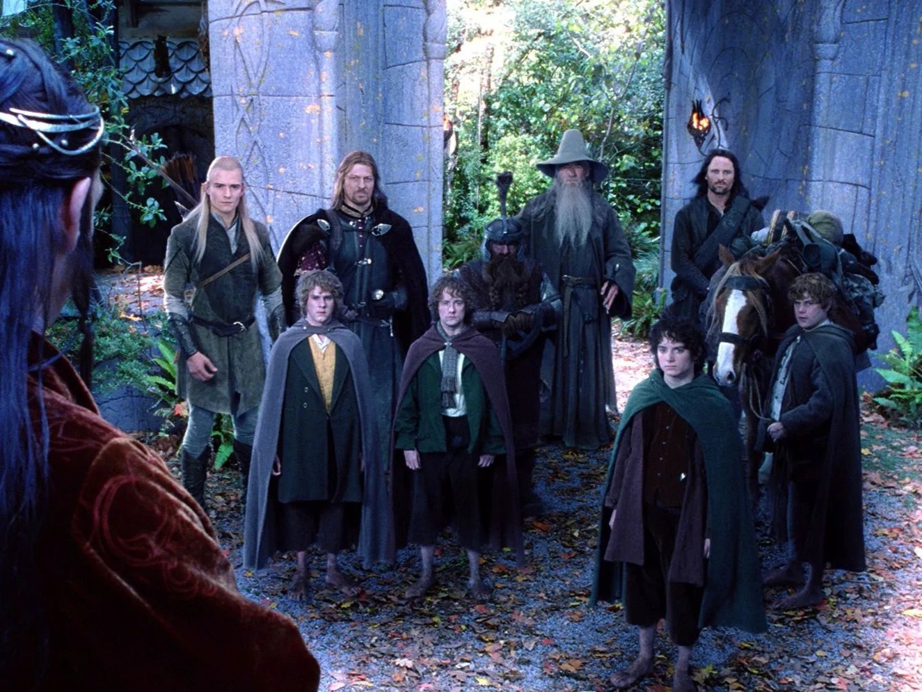 Personagens de O Senhor dos Anéis reunidos diante de uma construção de pedra em meio à floresta, usando trajes medievais; ao centro, um personagem de longas vestes e chapéu cinza segura um cajado, enquanto outros integrantes da comitiva aparecem ao redor.
